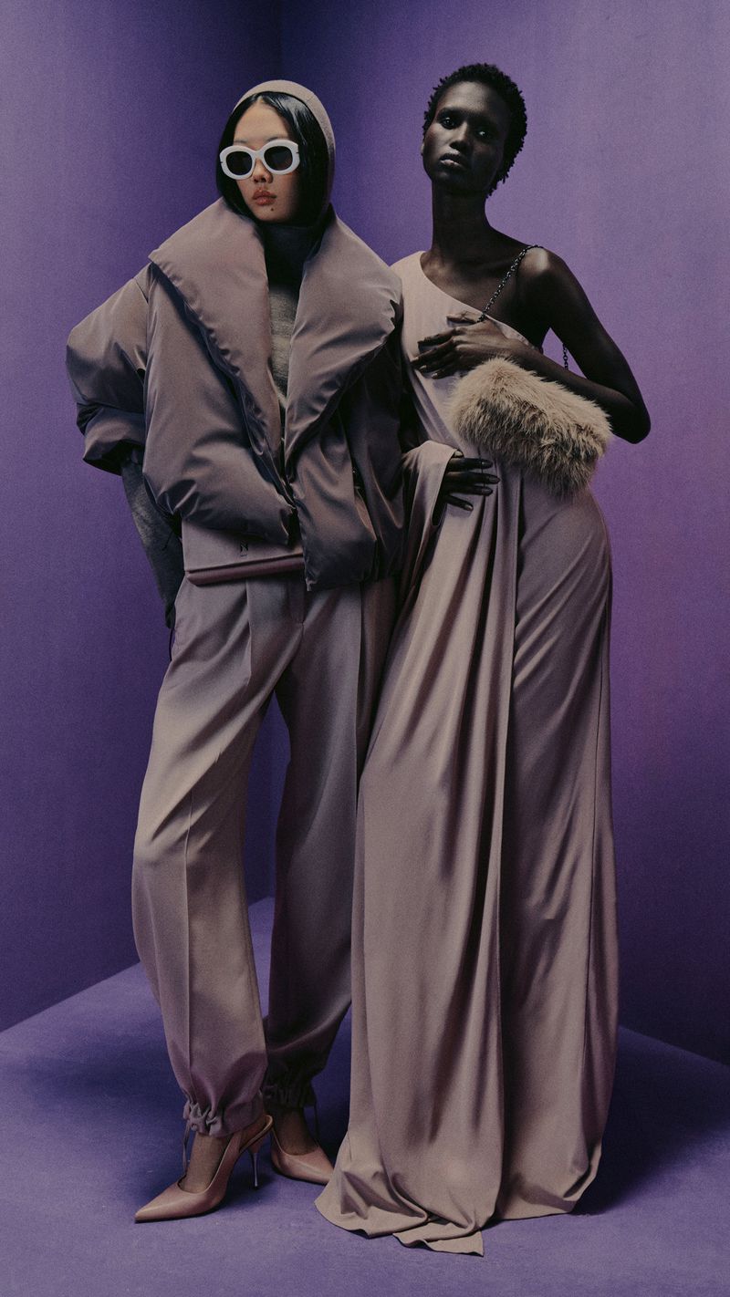 Two women in stylish purple and beige tones pose indoors; one in a puffer jacket and heels, the other in a draped dress with a fur muff.
