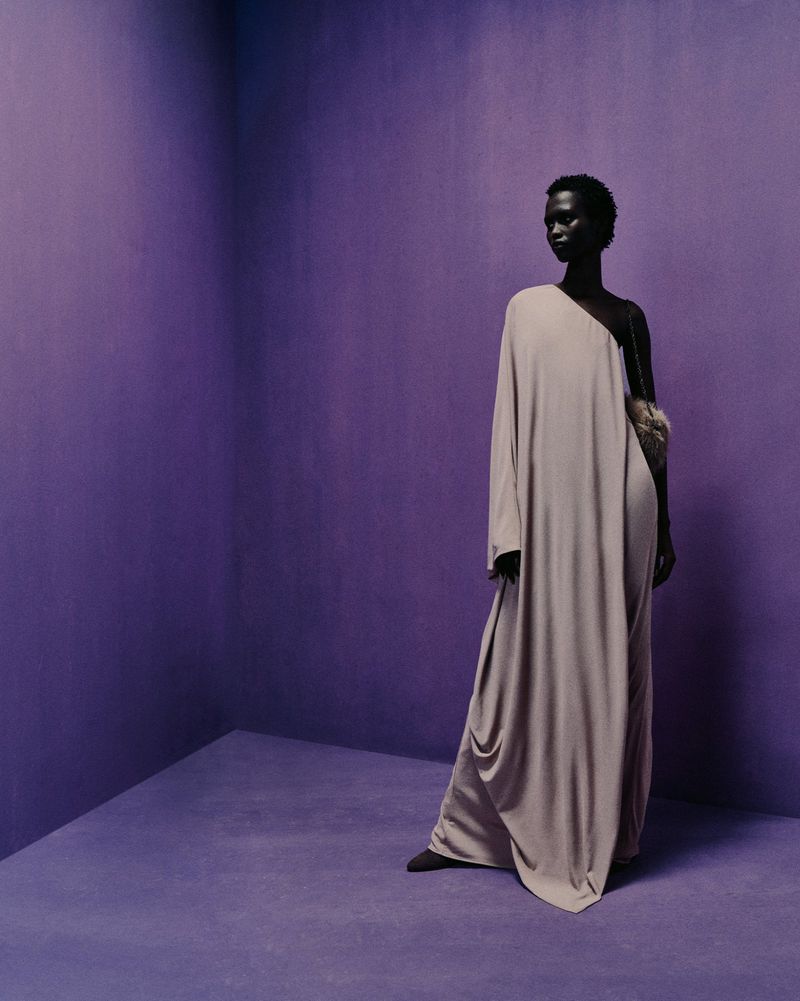A dark-skinned model poses in a lilac studio wearing a long, taupe gown with a furry handbag, creating a minimalist fashion photograph.