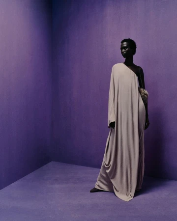 A dark-skinned model poses in a lilac studio wearing a long, taupe gown with a furry handbag, creating a minimalist fashion photograph.