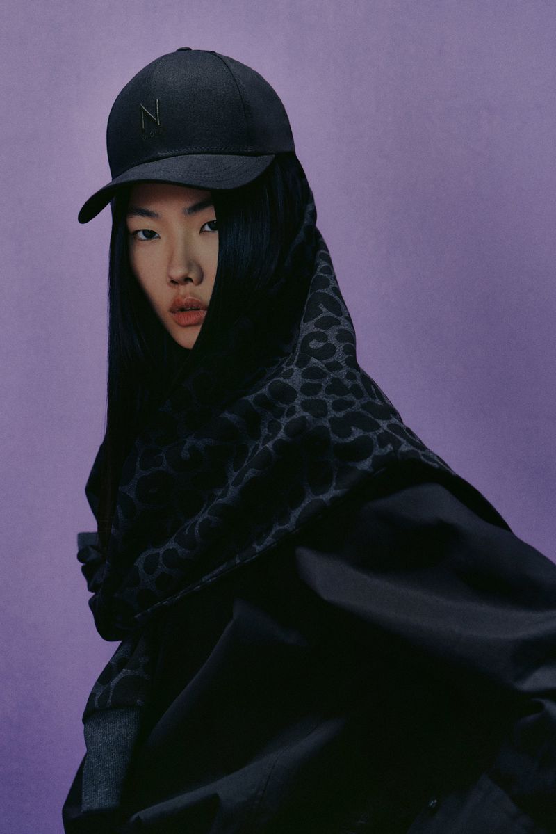 Asian adult female posing in black cap and patterned headscarf against a purple backdrop in a studio. Fashion editorial shot.