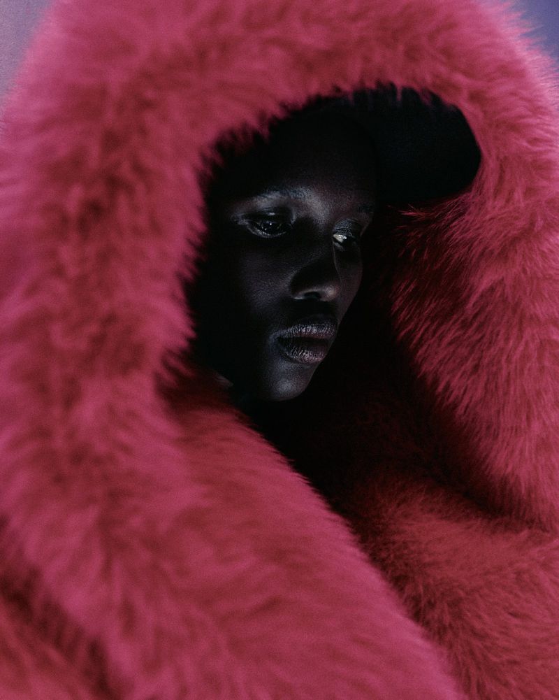Close-up portrait of a dark-skinned person wearing a bright pink faux fur hood, looking downwards with a serious facial expression.