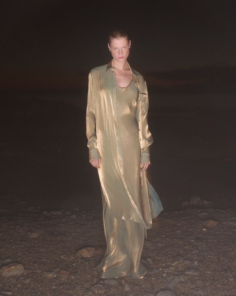 A woman stands on a barren landscape wearing a gold dress and shirt. The background is dark, with a small amount of sky visible.