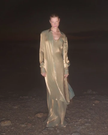 A woman stands on a barren landscape wearing a gold dress and shirt. The background is dark, with a small amount of sky visible.