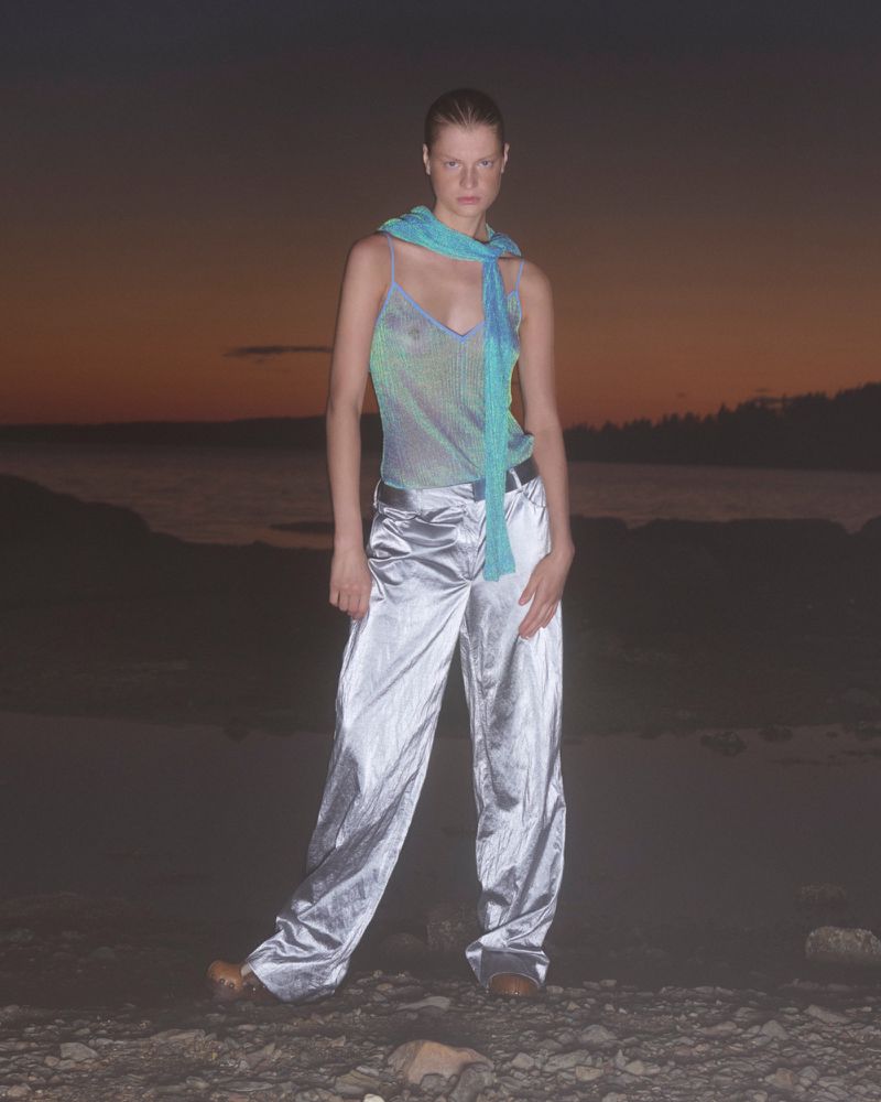 A woman stands outdoors wearing silver pants and a teal top against a dark dusk background, lit with dim natural light.