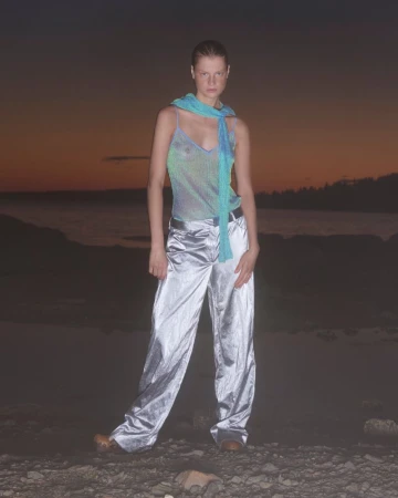 A woman stands outdoors wearing silver pants and a teal top against a dark dusk background, lit with dim natural light.