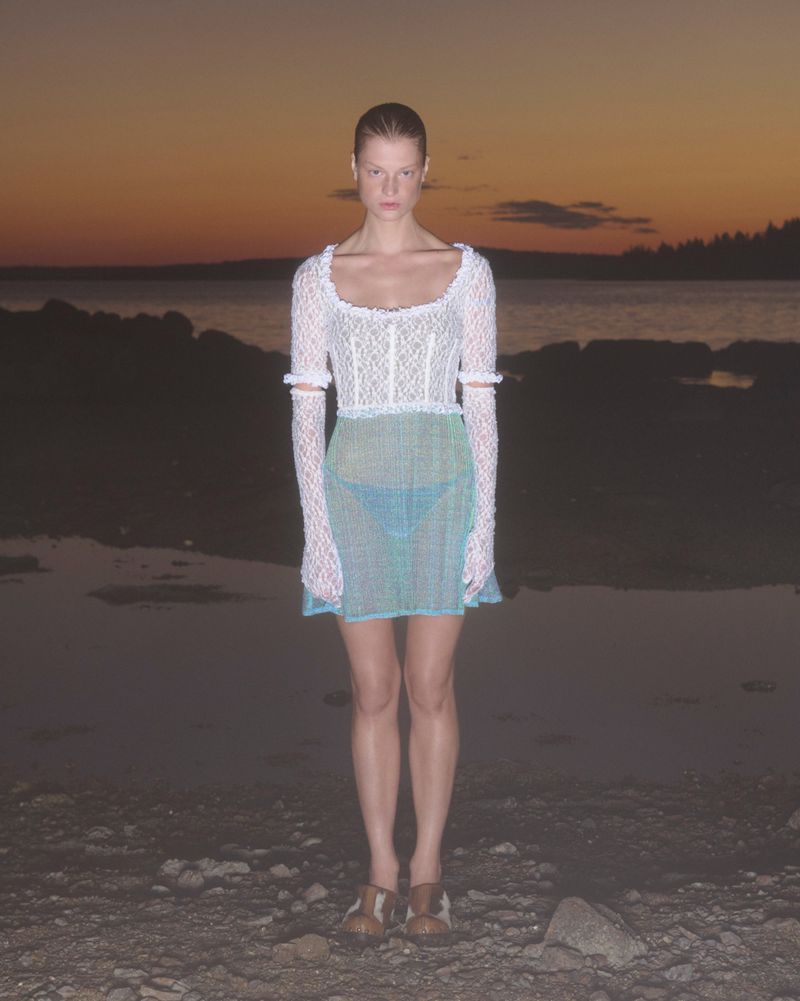 Woman in white lace top and teal skirt posing on rocky beach at sunset. Fashion shot with long sleeves and neutral expression.