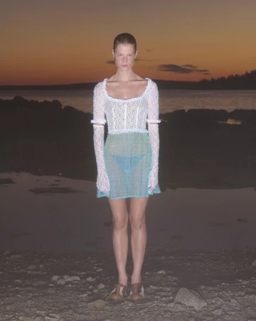 Woman in white lace top and teal skirt posing on rocky beach at sunset. Fashion shot with long sleeves and neutral expression.
