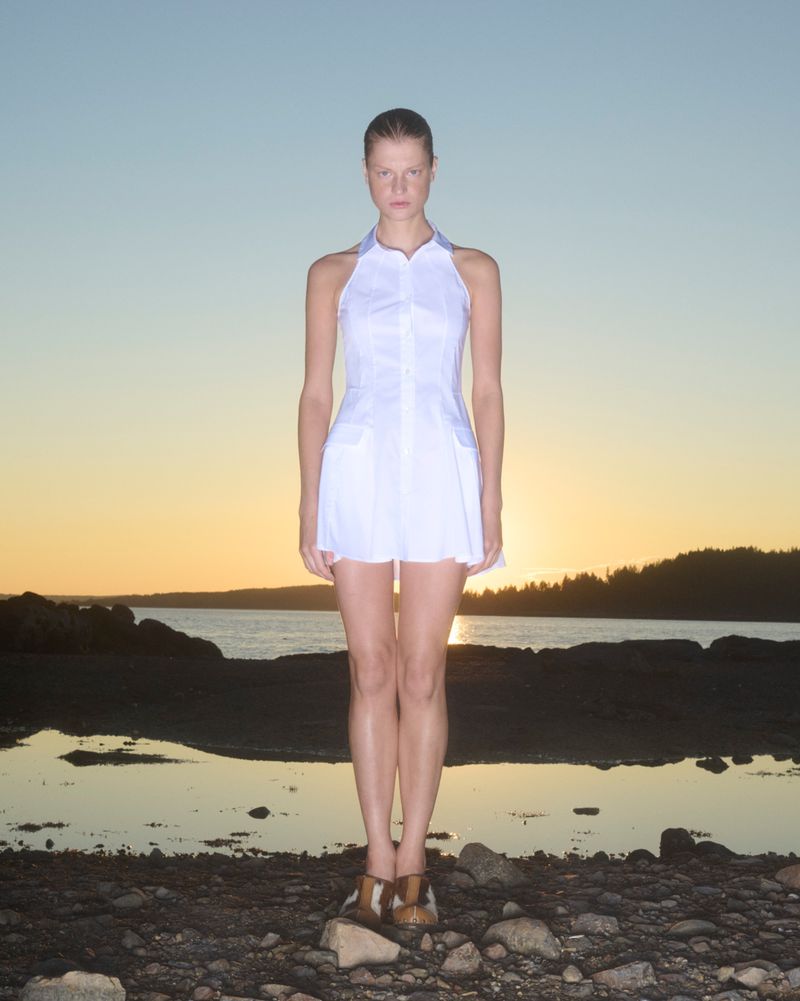 Woman in a white halter dress standing on rocks with a sunset over the ocean in the background, creating a bright and colorful horizon.