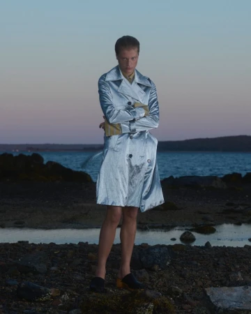 Adult wearing a shimmering silver trench coat and tan gloves, posing on rocky coastline with arms crossed at dusk with water backdrop.