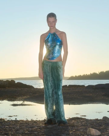 Woman in shimmering blue halter top and green velvet pants poses on a rocky beach at sunset, creating a fashion editorial image.