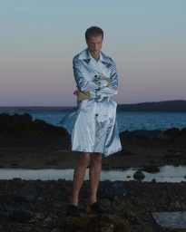 Model standing on a rocky shoreline wearing a metallic silver trench coat against an ocean and sunset sky background.