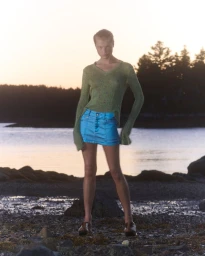 Model Michelle Laff poses on a rocky coast wearing a sheer olive green long-sleeve knit top and bright blue mini skirt.