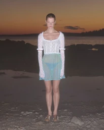 Model Michelle Laff wearing a white lace top with lace sleeves and a sheer blue mini skirt on a rocky beach at dusk.
