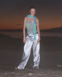 Model Michelle Laff posing in a sheer turquoise camisole and wide-leg metallic silver trousers on a rocky shore at dusk.