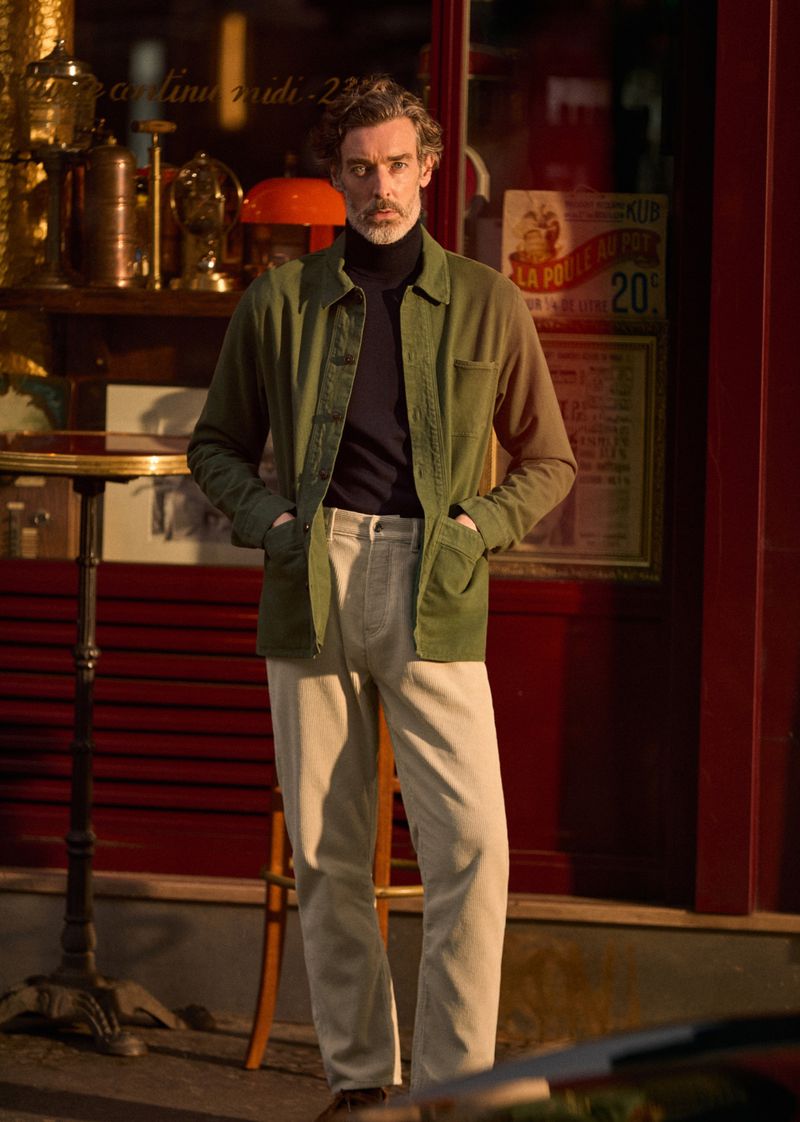 Full body shot of adult man in olive green jacket and beige pants standing outdoors in front of storefront window in daylight.