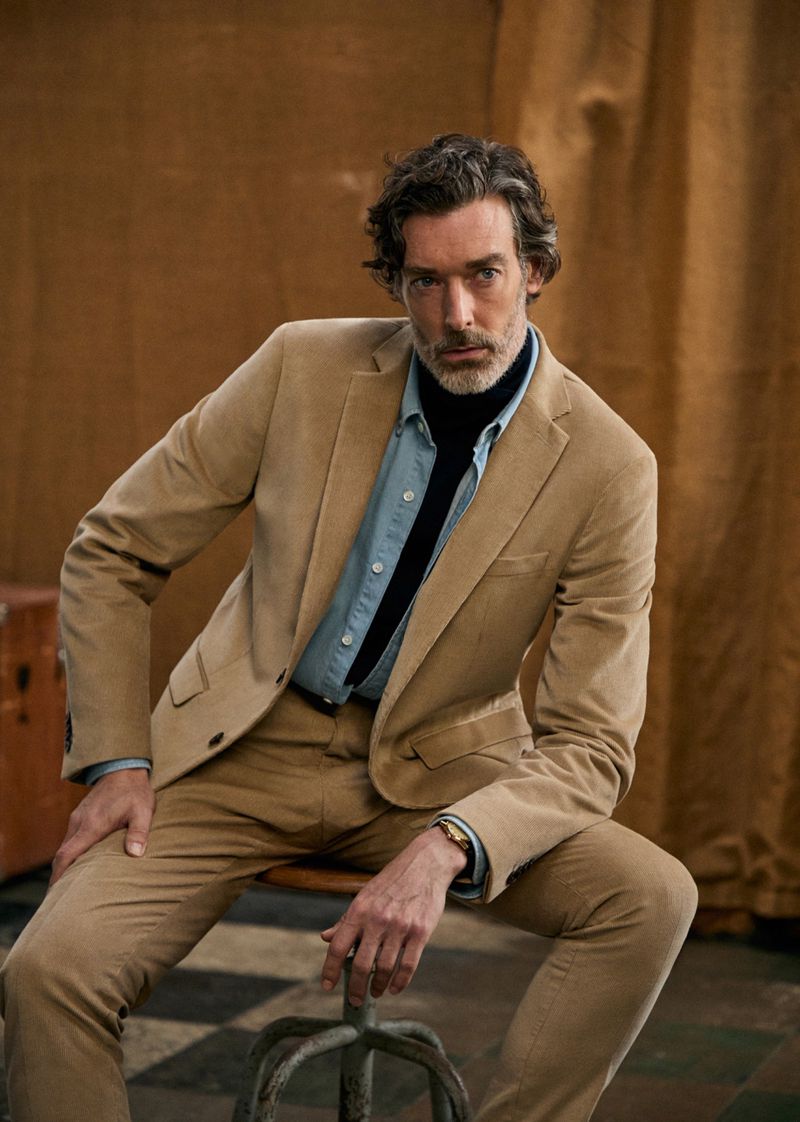 Portrait of adult male with beard and grey hair wearing a tan corduroy suit and light blue shirt while sitting on a metal stool.