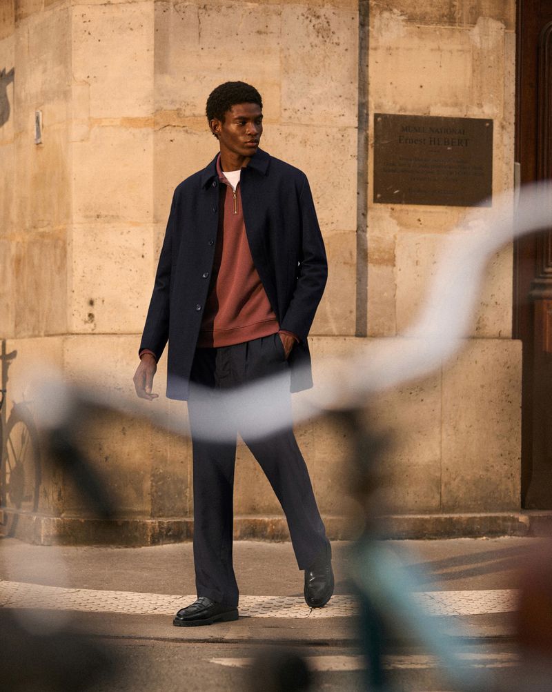 Stylish African-American man in dark coat and trousers poses against a stone wall in an urban setting, shallow depth of field.