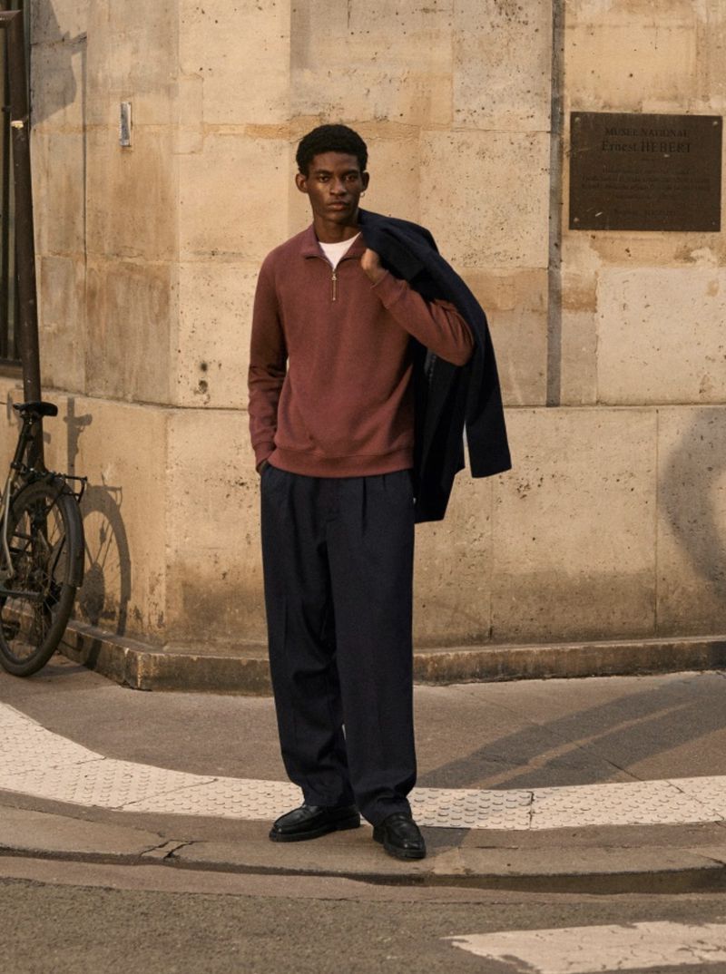 Adult male in a maroon pullover and navy pants stands on a sidewalk in an urban setting, holding a coat over his shoulder.