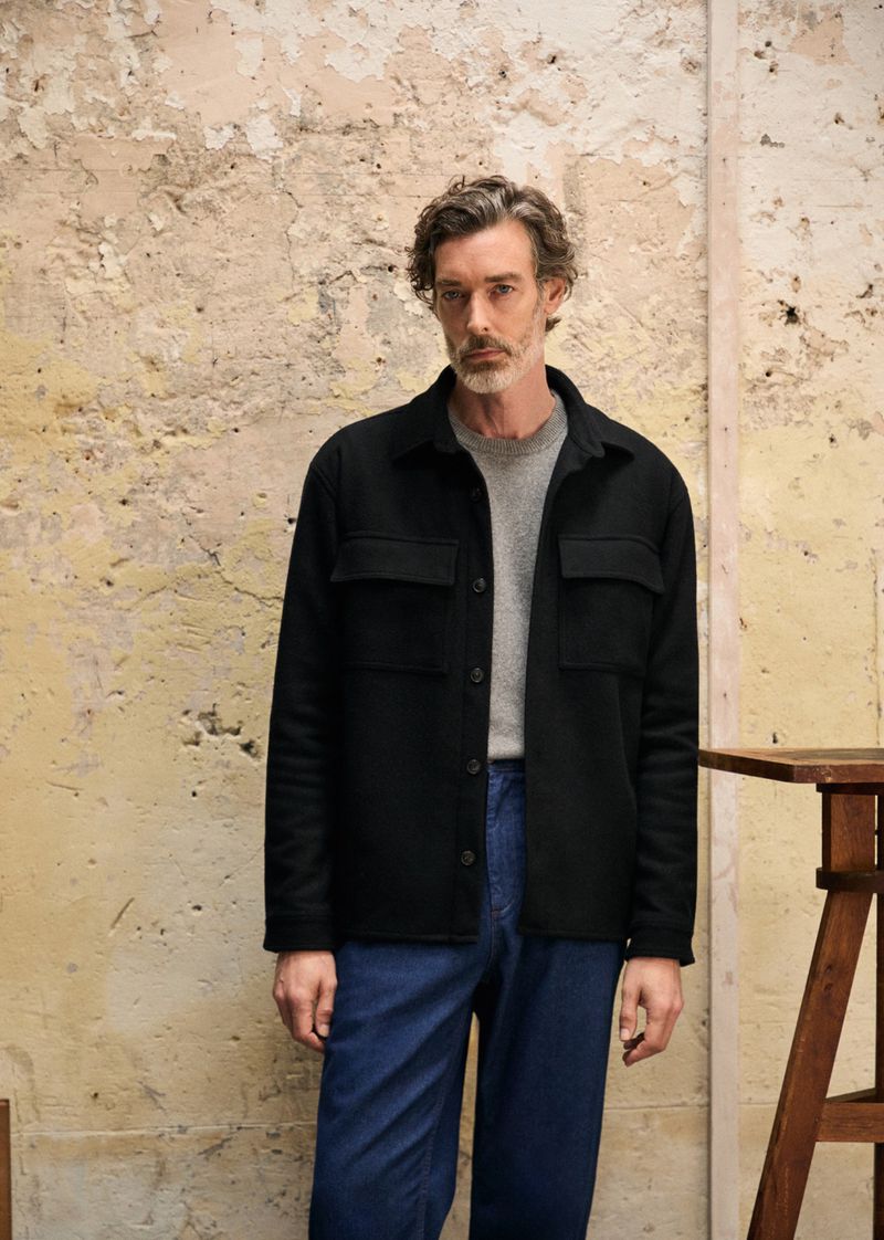 Man in a black button-down shirt poses in front of a textured wall, showcasing casual fashion and natural light photography.
