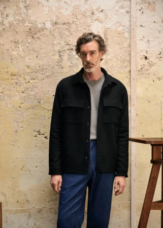 Man in a black button-down shirt poses in front of a textured wall, showcasing casual fashion and natural light photography.