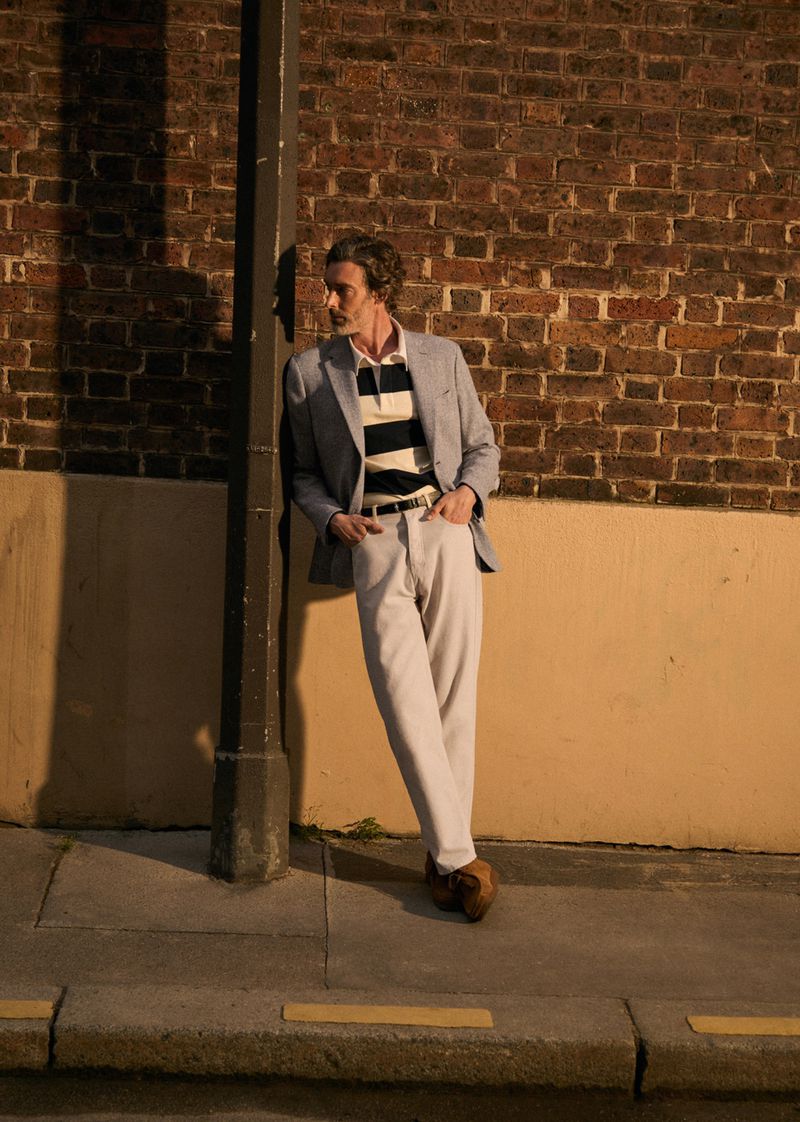 Man in gray blazer and white pants leaning against a post on a city street with a brick wall at night under dim, warm light.