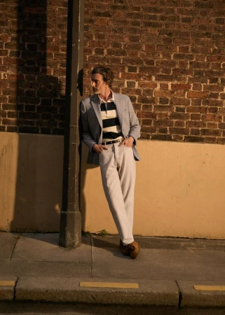 Man in gray blazer and white pants leaning against a post on a city street with a brick wall at night under dim, warm light.