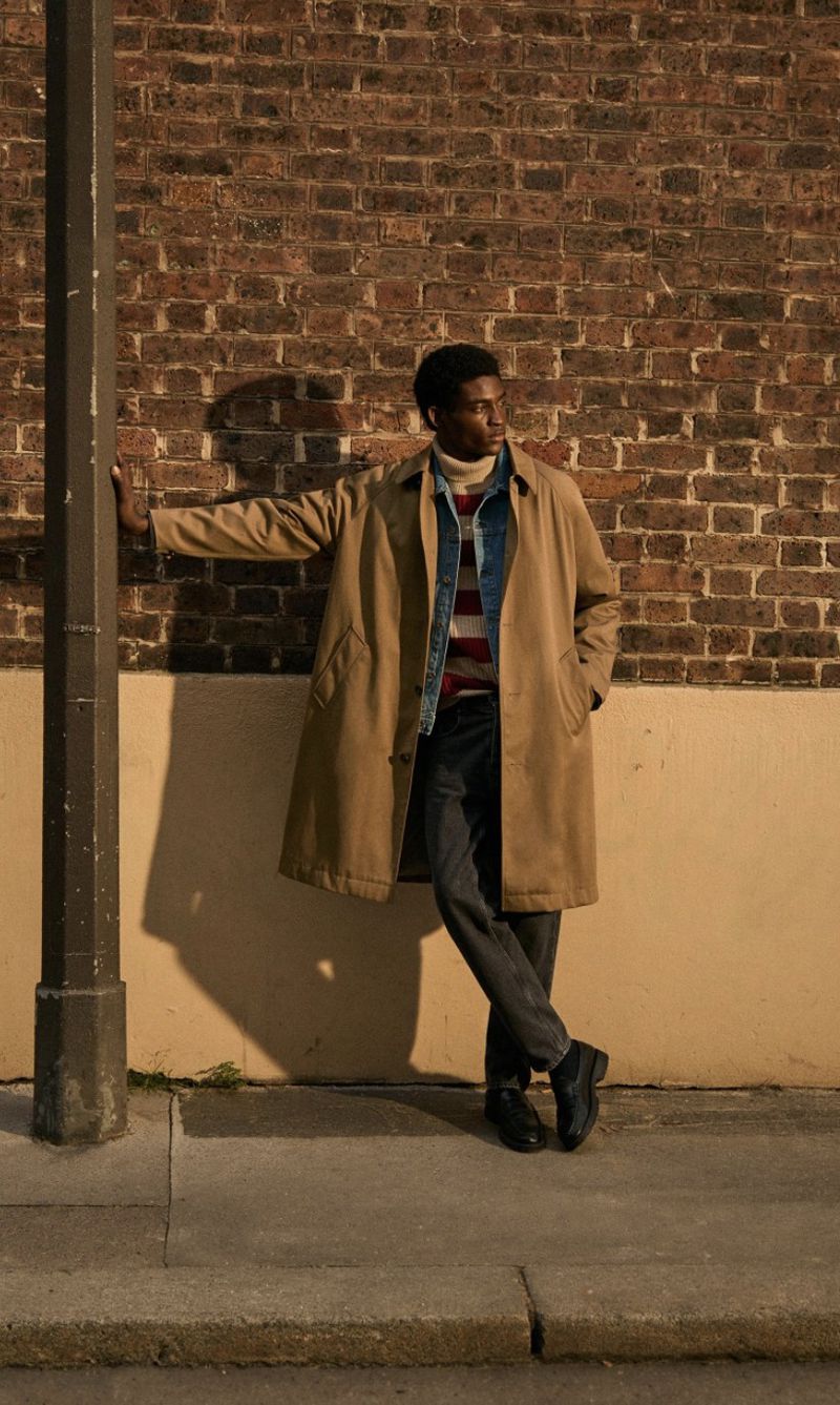 A Black male model in a beige coat leans against a pole near a brick wall. He wears a sweater, jeans, and black shoes, creating a fashionable urban scene.