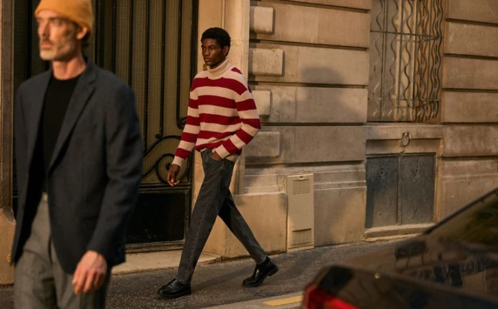 Two men walk on a city street; one in a red-striped sweater, the other in a blazer and beanie, with neutral stone buildings in the background.