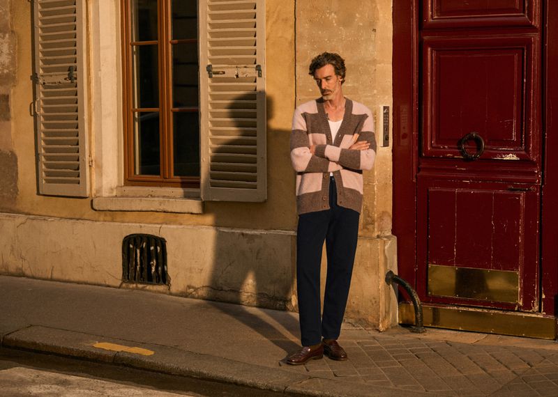 Adult male in cardigan and dark trousers stands in front of a red door and window with shutters on a brick paved street.