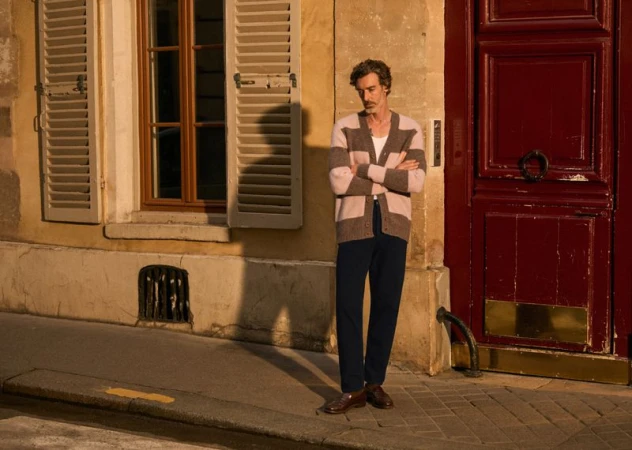 Adult male in cardigan and dark trousers stands in front of a red door and window with shutters on a brick paved street.