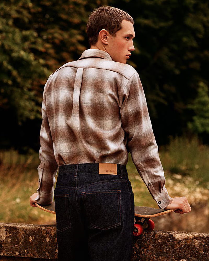 A young man stands outdoors, looking to the side while holding a skateboard, wearing a plaid shirt and dark denim jeans.