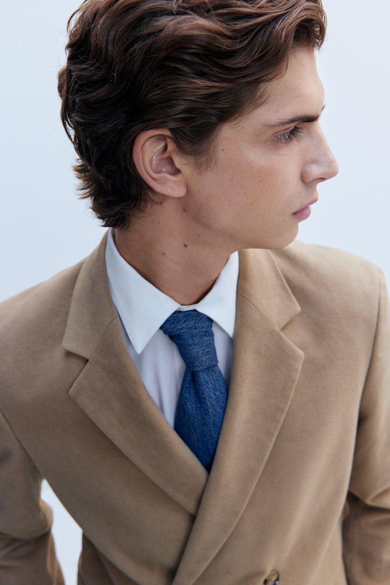 Close-up studio shot of an adult male with wavy brunette hair in a beige suit jacket, white shirt, and blue tie against a white backdrop.