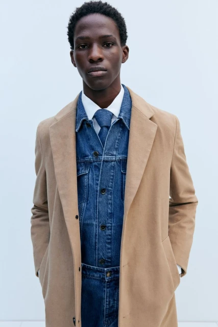 Studio shot of a Black male fashion model in a white collared shirt, denim jacket and pants, and tan coat in a neutral setting.