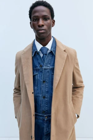 Studio shot of a Black male fashion model in a white collared shirt, denim jacket and pants, and tan coat in a neutral setting.