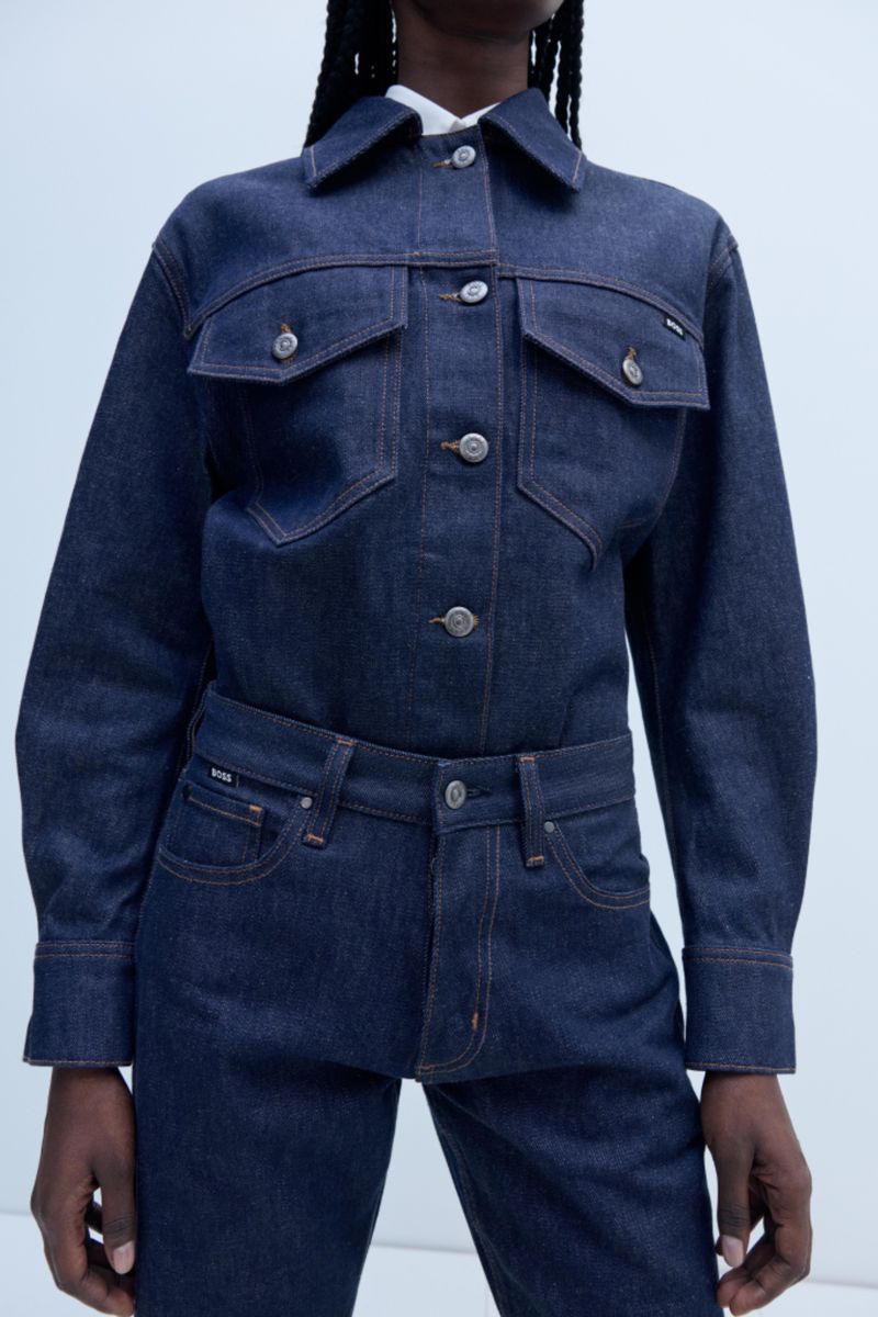 A medium studio shot presents an African-descent woman in a blue denim jacket and jeans, with a light background, showcasing fashion and style.
