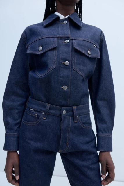 A medium studio shot presents an African-descent woman in a blue denim jacket and jeans, with a light background, showcasing fashion and style.