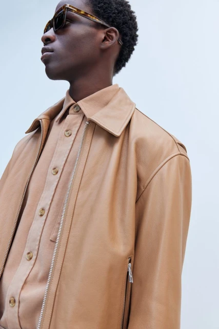 Profile view of a Black male model wearing a tan leather jacket and button-down shirt, complemented by sunglasses against a light background.