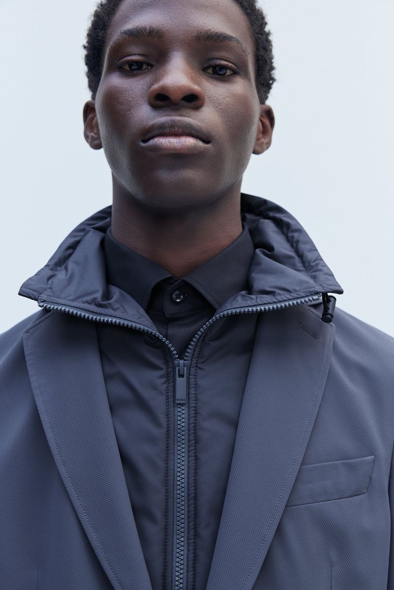 Close-up studio portrait of a Black male model wearing a black shirt, grey jacket, and puffy vest with a zipper. Neutral background.