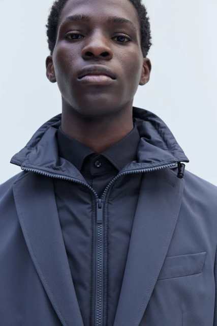 Close-up studio portrait of a Black male model wearing a black shirt, grey jacket, and puffy vest with a zipper. Neutral background.