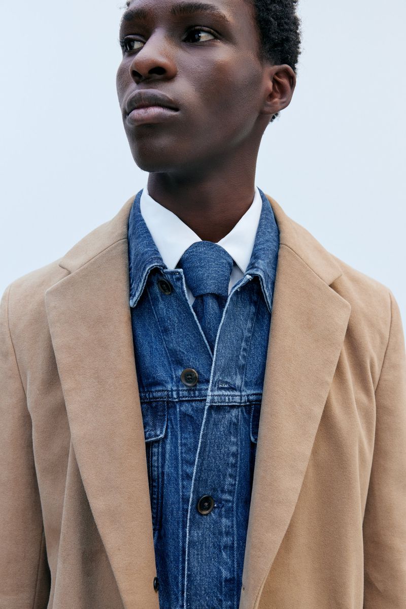Close-up of a Black man in a tan coat, denim jacket and tie over a white shirt, looking off-camera against a plain neutral background.