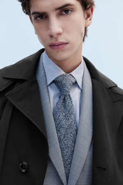 Close-up of a young adult man in a business-casual outfit with a trench coat, tie, striped shirt, and suit vest against a light-blue background.