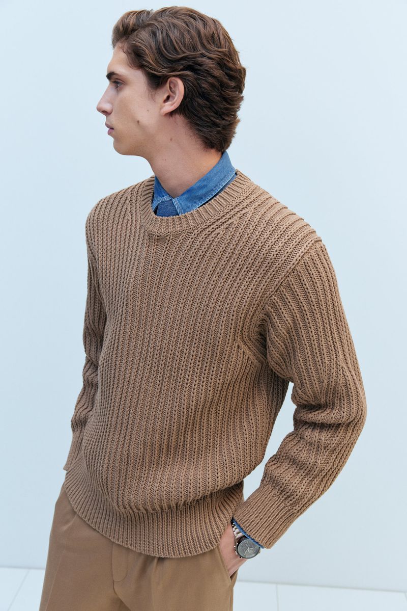 Profile shot of a light-skinned male model with wavy brown hair, wearing a tan sweater and pants, against a pale blue background.