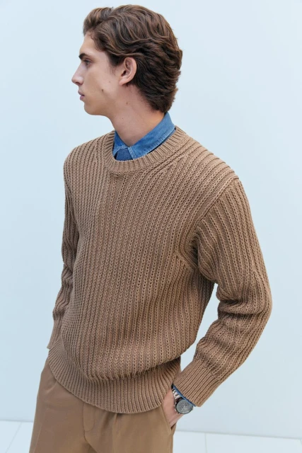 Profile shot of a light-skinned male model with wavy brown hair, wearing a tan sweater and pants, against a pale blue background.