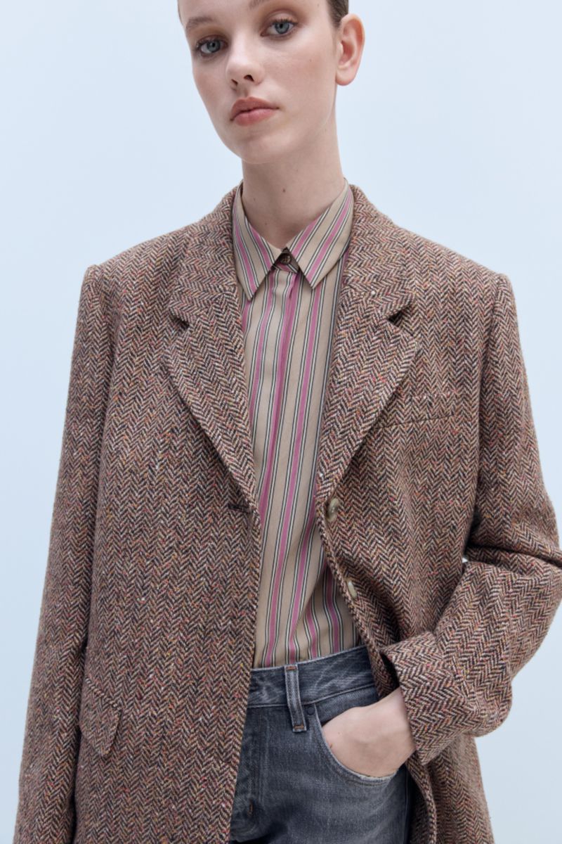 Close-up of a woman wearing a brown herringbone blazer and striped shirt against a light blue backdrop in an indoor studio setting.