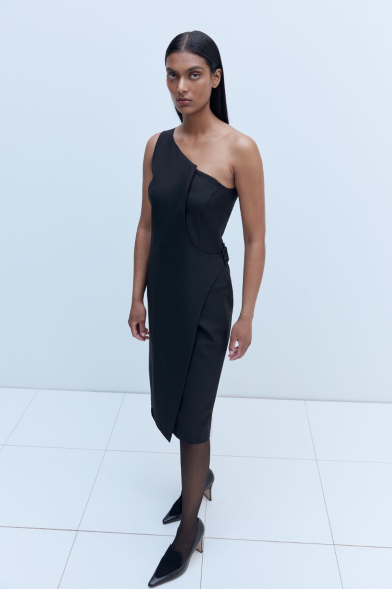 Elegant woman in a black, one-shoulder dress and black pointed heels, striking a pose against a clean, light-colored backdrop. 