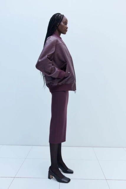 Profile shot of a Black female model wearing a burgundy bomber jacket and pencil skirt with black stockings in a studio setting.