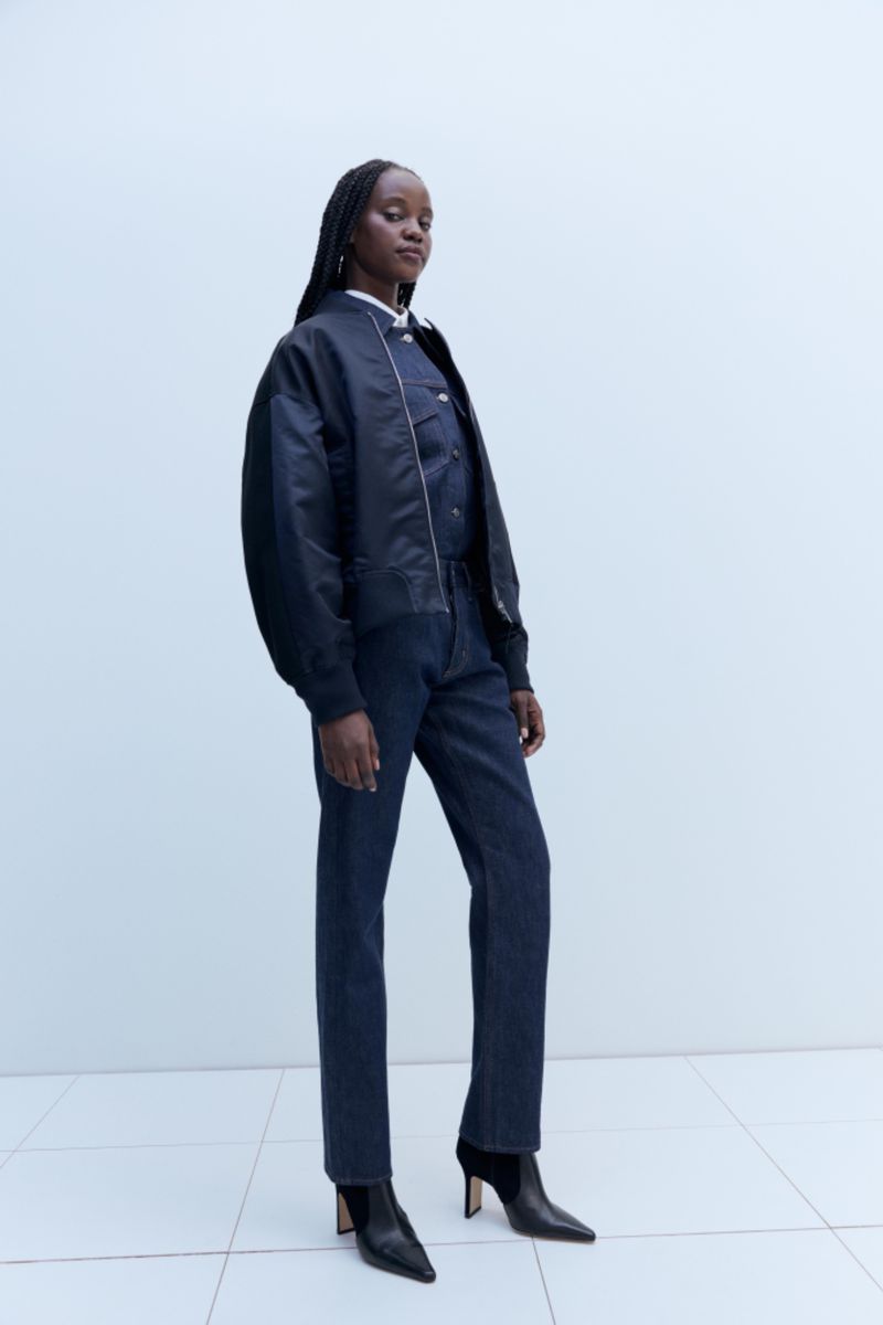 A Black woman is posing in a fashion shot wearing jeans, a bomber jacket, and boots against a pale blue background in a studio.