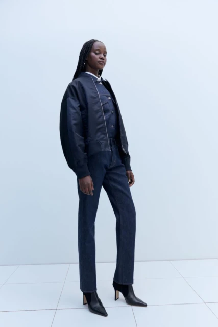 A Black woman is posing in a fashion shot wearing jeans, a bomber jacket, and boots against a pale blue background in a studio.