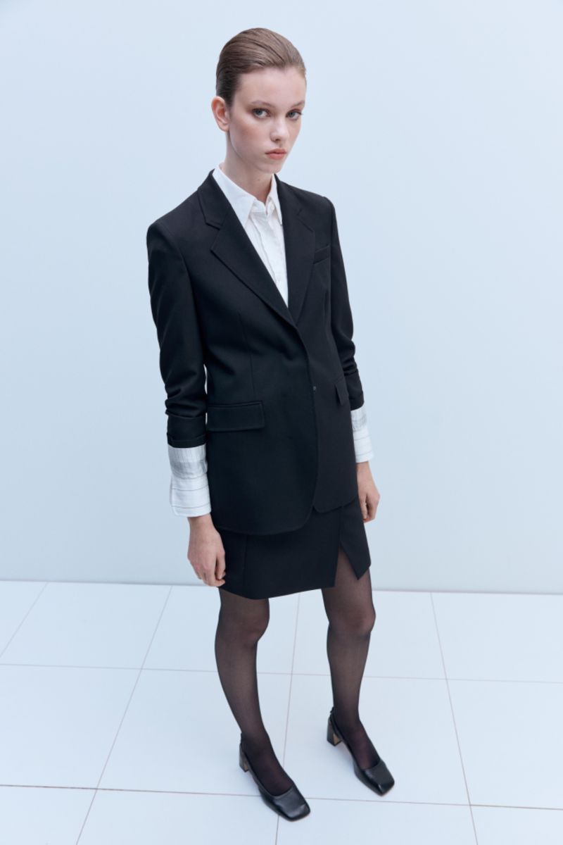 Model posing in a black tailored blazer, white collared shirt, matching black mini skirt, and sheer black tights against a pale studio wall.