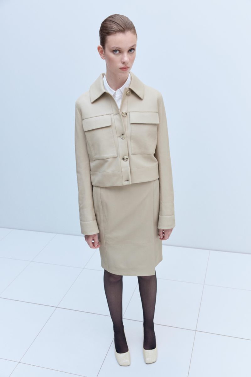 A fashionable adult woman is posing in a beige jacket and skirt outfit with black stockings and matching shoes on a white tile floor.
